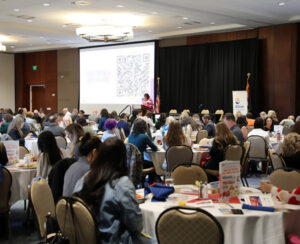 Conference room full of attendees with Liz Archuleta speaking at the podium and a QR code on the projected screen