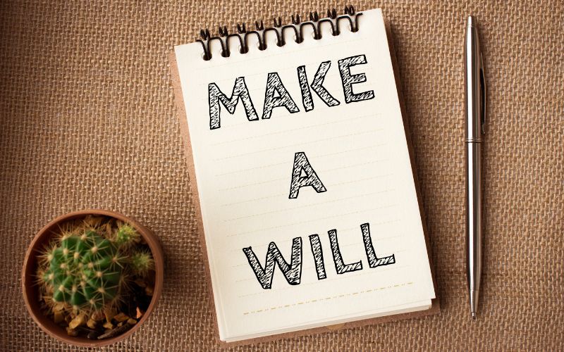 Pad of paper, pen and cactus plant on a table covered in burlap. On the pad it says Make A Will